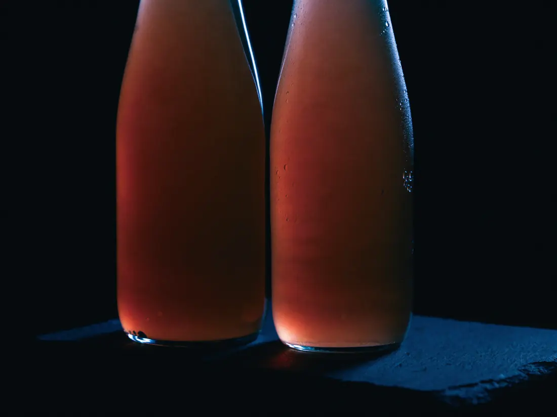 Two unlabelled glass bottles of infused beverage, beaded with condensation, on dark slate